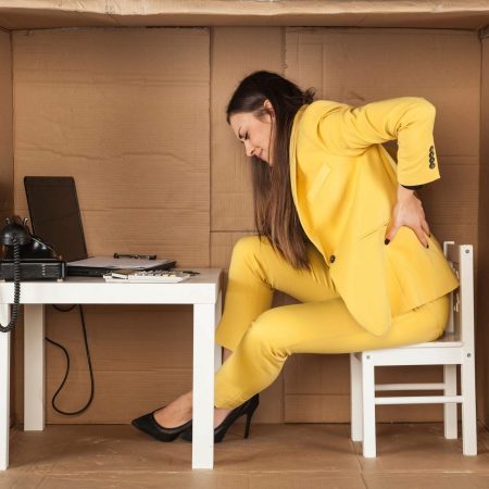 Canva – back pain cramped office and lack of space@2x Woman with back pain sitting at a desk set up in a cardboard box